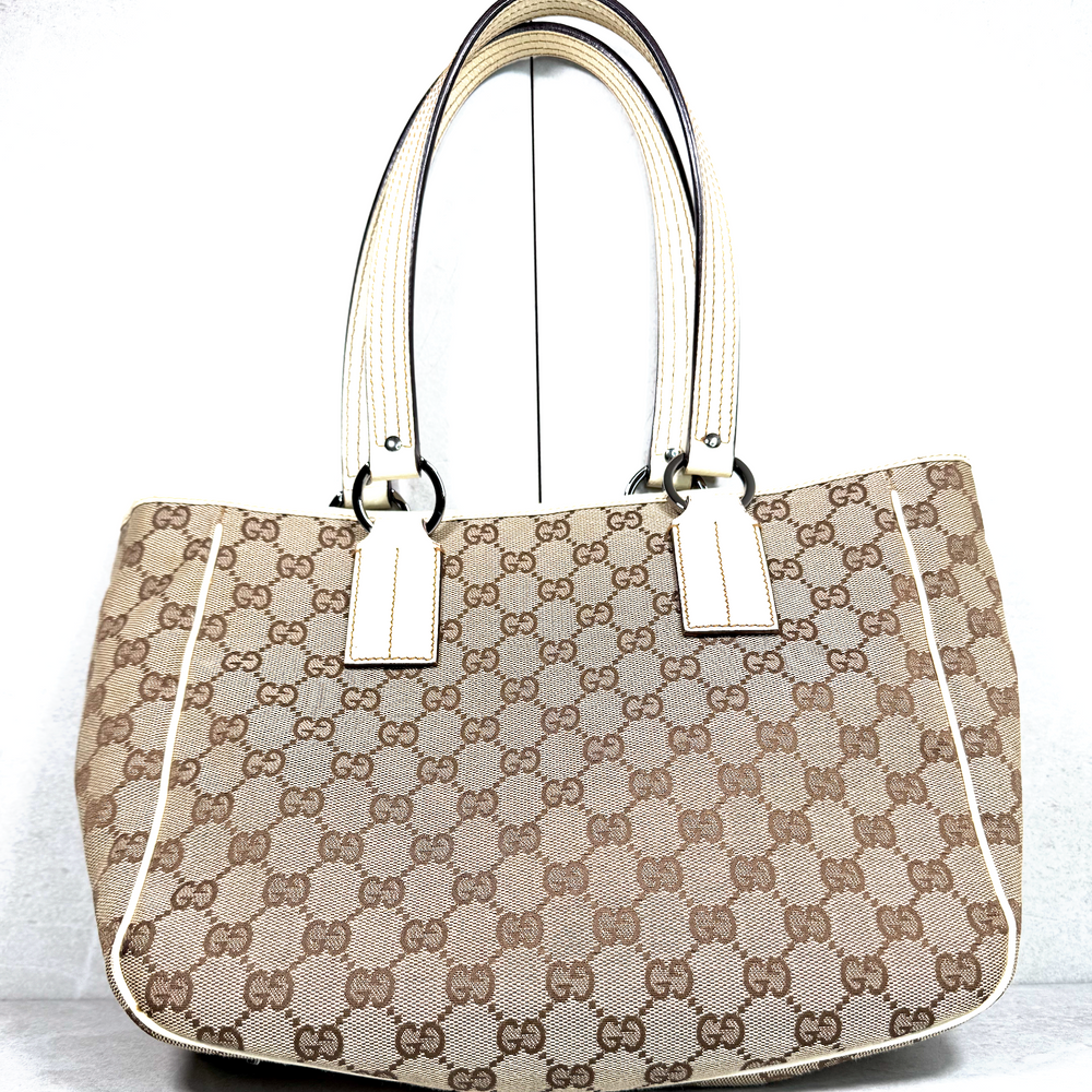 A Gucci tote bag in the classic brown and beige GG monogram canvas, featuring cream-colored leather handles and trim, photographed against a white background.
