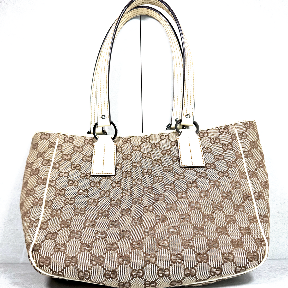 A Gucci tote bag in the classic brown and beige GG monogram canvas, featuring cream-colored leather handles and trim, photographed against a white background.