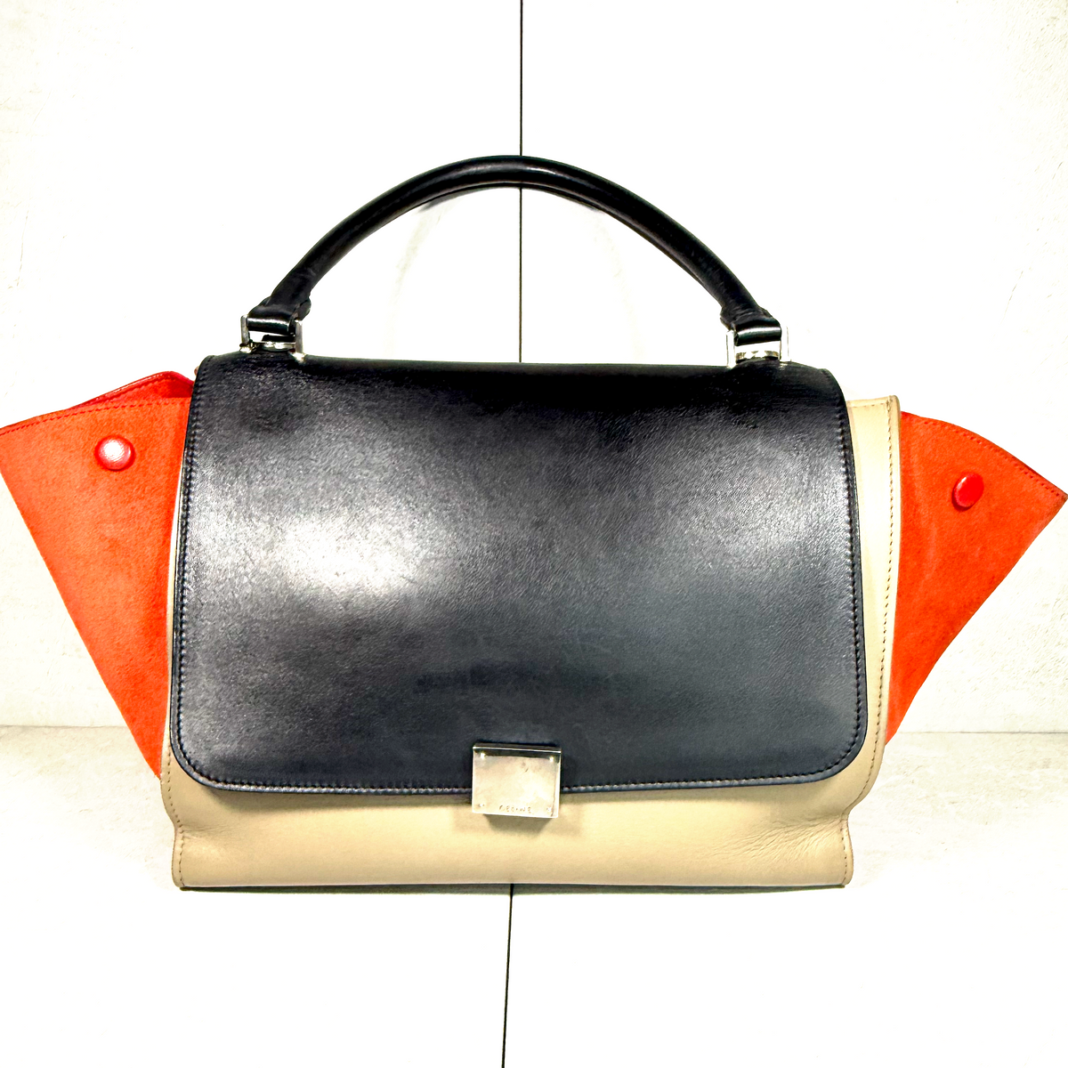 A structured leather handbag with a black top handle, a black front flap, a beige lower body, and bright orange side panels with red snap details, shown against a plain white background.